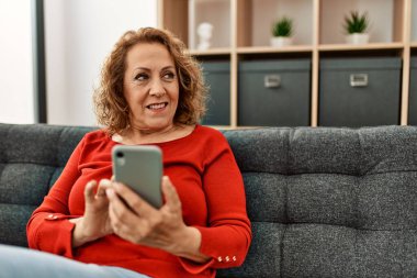 Orta yaşlı beyaz kadın akıllı telefon kullanıyor. Evdeki kanepede oturuyor..