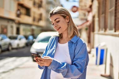 Genç sarışın kız şehirde akıllı telefon kullanarak mutlu bir şekilde gülümsüyor..