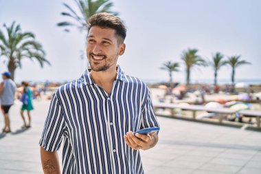 Genç yakışıklı adam deniz kenarında akıllı telefon kullanıyor.