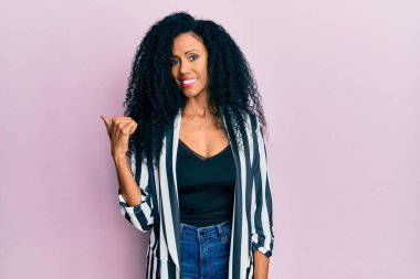 Orta yaşlı Afro-Amerikalı kadın, günlük kıyafetler giyiyor, gülümsüyor, gülümsüyor ve başparmağıyla yüzünü gösteriyor.. 