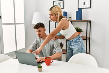 Genç beyaz çift dizüstü bilgisayar kullanıyor ve kahve içiyor. Evdeki masada oturuyorlar..