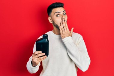 Sakallı, elinde motor yağı şişesi olan, ağzı elle kaplanmış, şok olmuş ve hata yapmaktan korkan genç bir İspanyol. şaşırmış ifade 