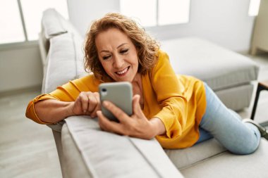 Orta yaşlı beyaz kadın akıllı telefon kullanıyor. Evdeki kanepede oturuyor..