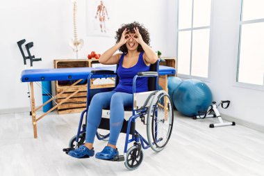 Orta Doğulu genç bir kadın, fizyoterapi kliniğinde tekerlekli sandalyede oturmuş dürbünün dili dışarı çıkarması, gözlerin parmaklarından dışarı bakması gibi güzel hareketler yapıyor. çılgın ifade. 