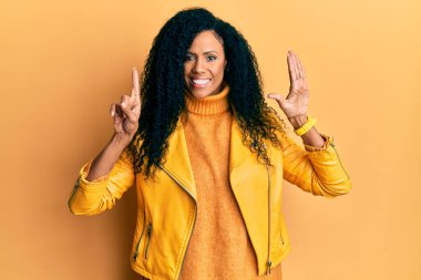 Orta yaşlı Afro-Amerikalı kadın yün kazak ve deri ceket giyip altı numaralı parmaklarını gösterirken kendinden emin ve mutlu bir şekilde gülümsüyor.. 