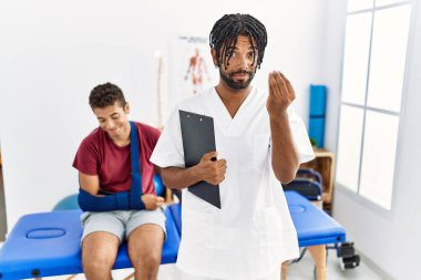 Genç İspanyol bir adam kırık kollu bir adamla birlikte ağrı kesici kliniğinde çalışıyor. El ve parmakları kendinden emin bir ifadeyle italyan hareketi yapıyor. 