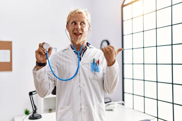 身穿医生制服的金发年轻人拿着听诊器站在诊所 指尖着大拇指朝边笑着 高兴地张开嘴 — 图库照片