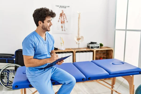 Young Hispanic Man Wearing Physio Therapist Uniform Writing Clipboard Clinic — Stockfoto