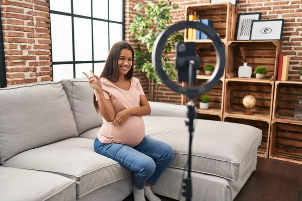 若い妊婦は勝利の印をするカメラで幸せな顔のウィンクで笑顔で自宅でスマートフォンでVlogチュートリアルを記録します — ストック写真