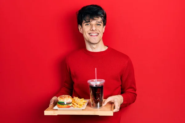Handsome hipster young man eating a tasty classic burger with fries and soda smiling with a happy and cool smile on face. showing teeth.