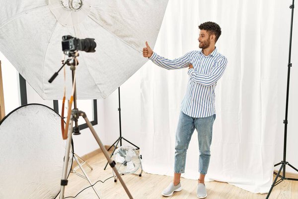 Arab young man posing as model at photography studio looking proud, smiling doing thumbs up gesture to the side 