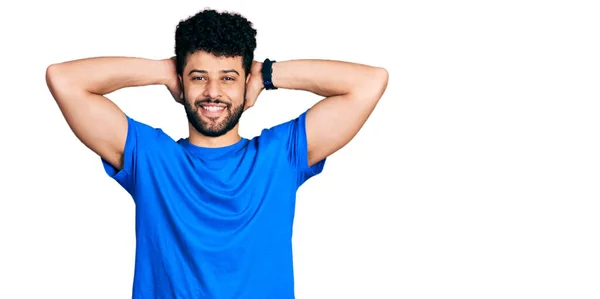 年轻的阿拉伯男子 留着胡须 穿着休闲的蓝色T恤 放松和伸展 胳膊和手背在头和脖子后面开心地笑着 — 图库照片