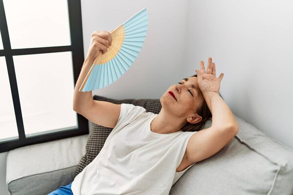 Латиноамериканка средних лет улыбается сидя на диване, используя ручной вентилятор дома