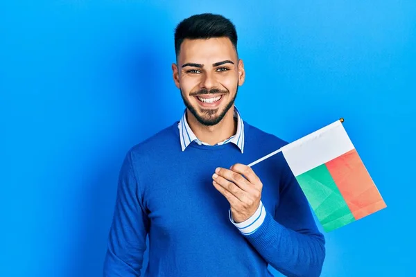 Joven Hombre Hispano Con Barba Sosteniendo Bandera Madagascar Luciendo Positivo —  Fotos de Stock