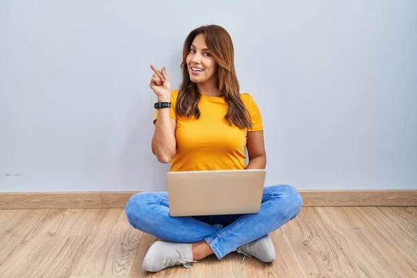 西班牙女人用笔记本电脑坐在家里的地板上 脸上挂着大大的笑容 手指头指向旁边看相机 — 图库照片