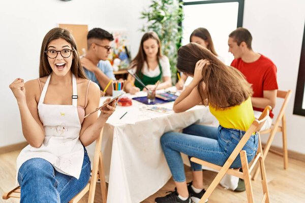 Молодая художница рисует в художественной студии, гордо крича, празднуя победу и успех, очень взволнованная поднятыми руками 