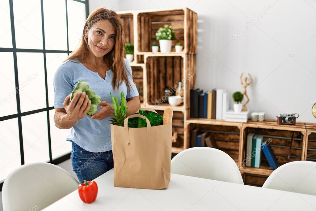 Middle age caucasian woman holding paper bag with groceries standing at home.