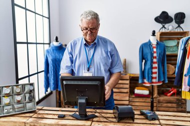 Orta yaşlı, gri saçlı, gülümseyen, kendine güvenen bir adam giyim mağazasında çalışıyor.