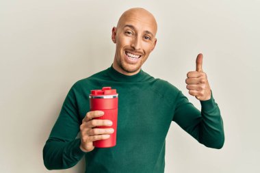 Kel ve sakallı bir adam bir fincan kahve içiyor mutlu ve pozitif gülümsüyor, başparmağını kaldırıyor ve işaret veriyor. 