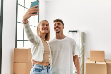 Genç beyaz çift mutlu bir şekilde gülümsüyor. Yeni evlerinde akıllı telefonların yanında selfie çekiyorlar..