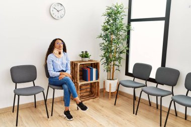 Klinik bekleme odasında sandalyeye oturan yaka takan genç Latin kadın.