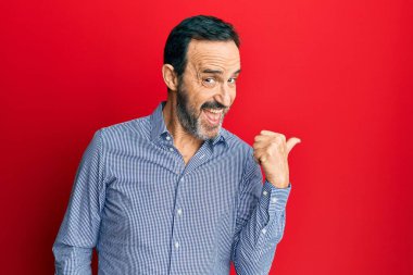 Orta yaşlı İspanyol bir adam, günlük kıyafetler giyiyor, gülümsüyor, gülümsüyor ve başparmağıyla yan tarafa bakıyor.. 