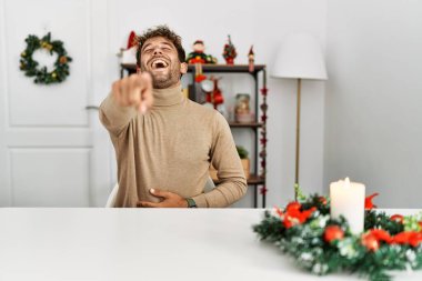Masada sakallı genç yakışıklı adam noel süslemesinin yanında sana gülüyor, eli vücudunun üzerinde kamerayı işaret ediyor, utanç ifadesi takınıyor. 