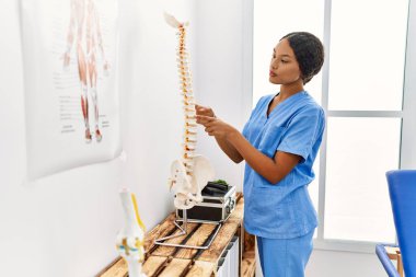 Fizyoterapist üniforması giyen genç Latin kadın klinikte parmağıyla omurgasını işaret ediyor.