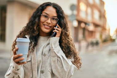 Genç Latin kadın akıllı telefondan konuşuyor ve şehirde kahve içiyor..