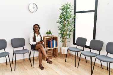 Genç Afrikalı Amerikalı kadın bekleme odasında akıllı telefon ve kulaklık kullanıyor..