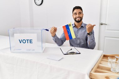 Siyasi seçim kampanyasında sakallı, genç yakışıklı bir adam elinde Venezuela bayrağı ile başparmağını kaldırıp ağzı açık gülümsüyor. 