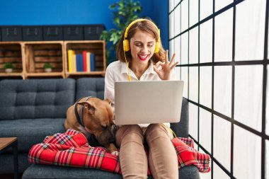 Genç ve güzel bir kadın kanepede dizüstü bilgisayar kullanıyor parmaklarıyla imza atıyor, gülümseyerek mükemmel bir sembol oluşturuyor. 