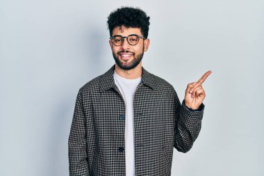 Sakallı, gözlüklü, yüzünde kocaman bir gülümseme olan, eli ve parmağı kameraya bakan genç bir Arap.. 