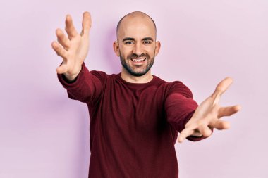 Sıradan kıyafetler giyen genç kel adam kameraya sarılıp gülümseyerek gülümsüyor. Mutluluğu kucaklayan neşeli bir ifade.. 