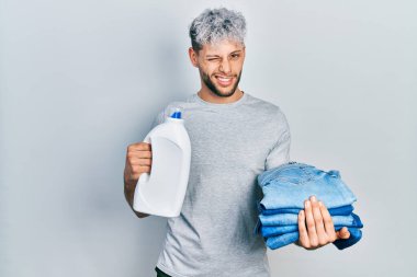 Modern boyalı saçlı, çamaşırhane için kot pantolon tutan ve deterjan şişesi tutan genç İspanyol bir adam kameraya seksi bir ifadeyle bakıyor, neşeli ve mutlu bir yüzü var.. 