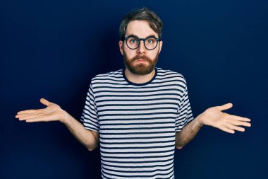 Çizgili tişört giyen, gözlüklü, kafası karışık ve elleri ve kolları havada beyaz bir adam. şüphe kavramı. 