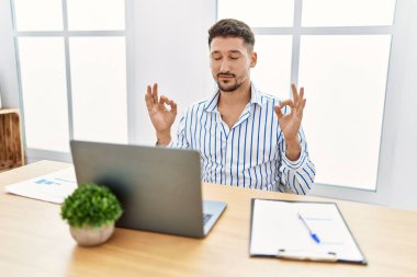 Ofiste sakallı genç yakışıklı adam bilgisayar kullanıyor rahatla ve gözleri kapalı gülümse parmaklarıyla meditasyon hareketi yapıyor. yoga konsepti. 