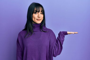 Kâküllü, balıkçı yaka kazaklı, neşeli gülümseyen, avuç içi kameraya bakan genç esmer kadın.. 