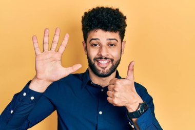 Sakallı, sıradan gömlekli genç bir Arap. Altıncı parmaklarını gösterip gösterirken kendinden emin ve mutlu bir şekilde gülümsüyor.. 
