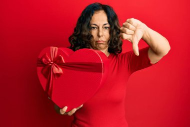 Orta yaşlı İspanyol kadın elinde kızgın suratlı sevgililer günü hediyesi tutuyor. Eksi işareti baş parmakları aşağıda. Reddedilme kavramı. 