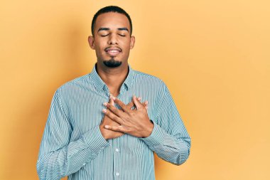 Sıradan kıyafetler giyen, göğsünde elleri kapalı, yüzünde minnet dolu jestlerle gülümseyen Afro-Amerikan bir genç. sağlık kavramı. 