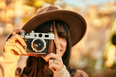 Kışlık şapkalı esmer kadın parkta klasik kamera kullanarak gülümsüyor.
