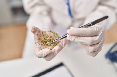 Laboratuvarda bilim adamı üniforması giyen genç Latin kadın elinde cımbızla bitki örneği tutuyor.