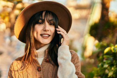 Kışlık şapkalı esmer kadın parkta telefonda gülümsüyor.