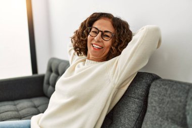 Orta yaşlı İspanyol kadın evde gözlük takarak gülümsüyor.