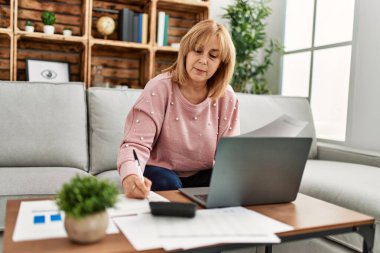 Orta yaşlı sarışın kadın evdeki kanepede oturarak tele-çalışmaya odaklanmış..