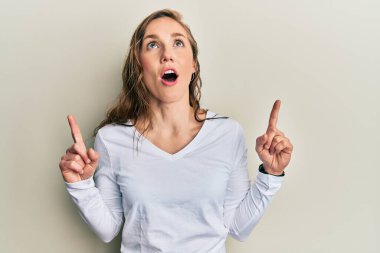 Gündelik kıyafetler giyen genç sarışın kadın şaşırmış ve şaşırmış bir şekilde yukarı bakıyor ve parmaklarıyla işaret ediyor ve kollarını kaldırıyor.. 