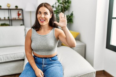 Evdeki kanepede oturan genç esmer kadın merhaba diyor mutlu ve gülümsüyor, dostça bir hoş geldin jesti. 