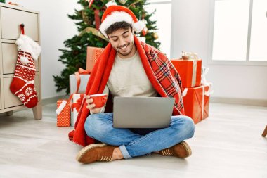 Genç İspanyol adam Noel şapkası takıyor dizüstü bilgisayar kullanıyor ve evde kahve içiyor..