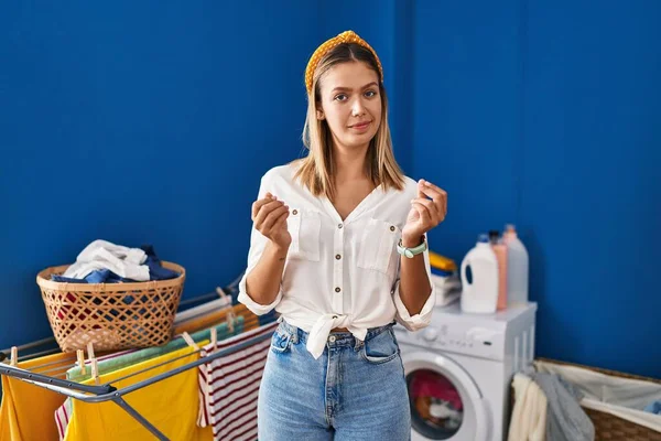 Young blonde woman at laundry room doing money gesture with hands, asking for salary payment, millionaire business